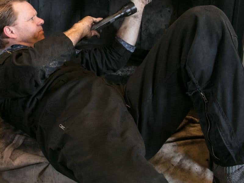 A chimney sweep cleaning a fireplace. He's wearing black overalls and shining a torch up the chimney to inspect it. He's lying on a dust sheet to protect the floor from the soot from the chimney.