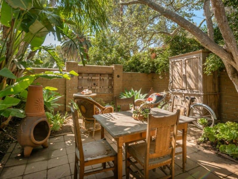 A secluded area in the back yard of a house with a dining table and chairs and a chimenea. The area is enclosed with an adobe wall and paved with stone slabs.