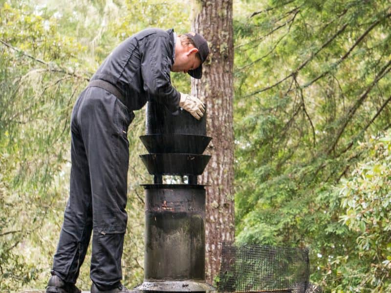 A construction worker wearing black overalls installs a precast metal chimney for an adobe fireplace. He stands next to the top of the chimney on the roof of the house, looking down it.