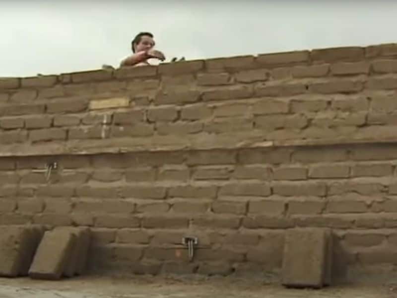 A screenshot of a video by Building and DIY on YouTube. A worker is laying an adobe wall. His head is visible over the top of the wall that he's working on the other side of. Adobe bricks are leaning against the front of the wall.