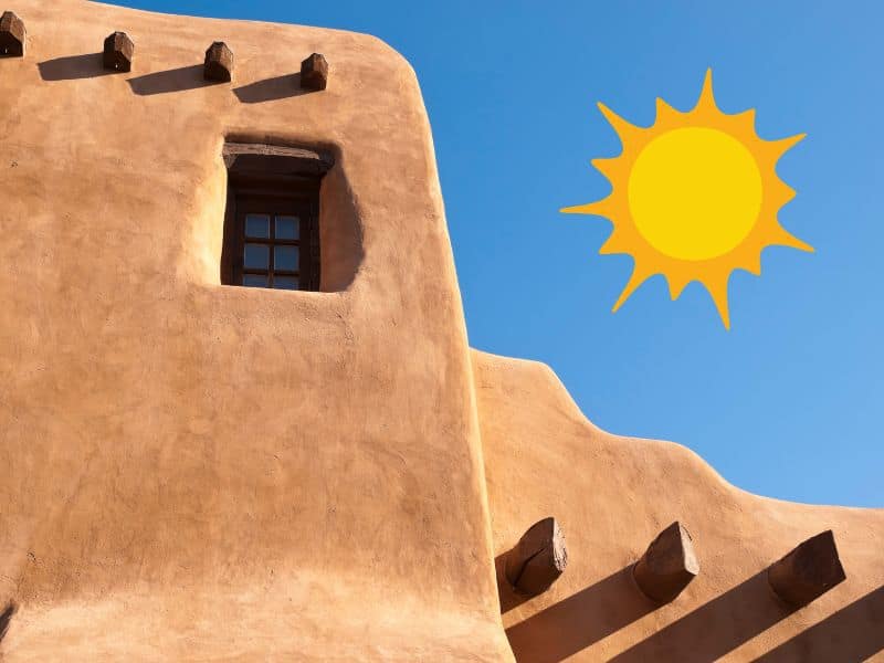 An orange adobe house with small windows and wooden beams protruding from the wall, which cast shadows along the wall. To the right is a bright sun shining.