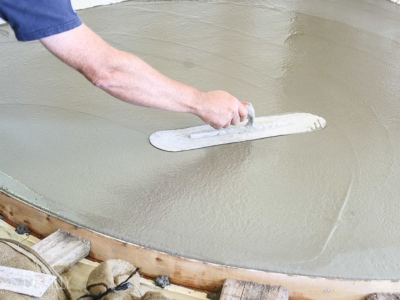 A worker is preparing the foundation for the hearth by smoothing the concrete with a float. Wooden forms can be seen in the bottom of the photograph, which contain the concrete and give it the right shape.