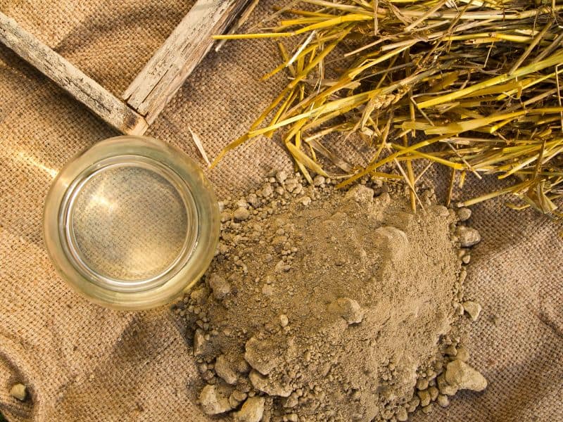 A photograph of piles of straw and dirt and a glass of water beside a wooden form for making adobe bricks.