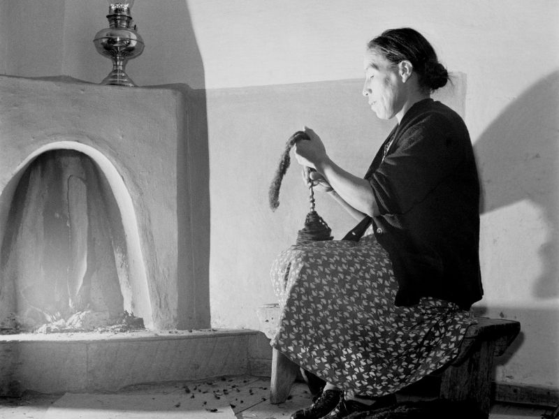 An old black and white photograph of an adobe fireplace with an old woman spinning yarn in front of it but the light of the fire and an oil lamp resting on the mantelpiece.