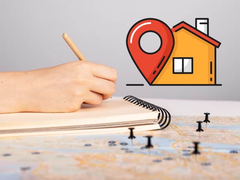 A photograph of a person's hand writing in a spiral-bound notebook resting on a map with 5 pins in it. In the top right is an icon of a house with a location pin alongside.