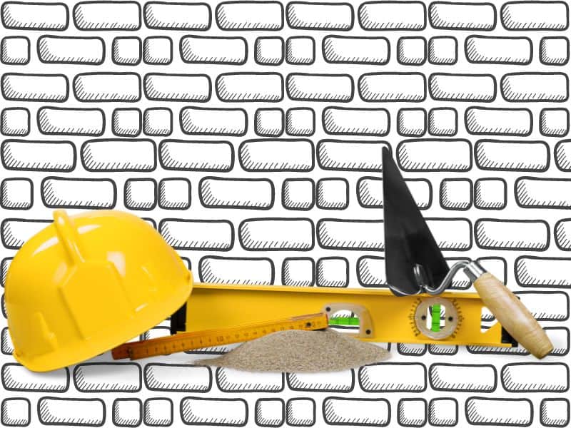 Masonry tools including a yellow hard hat, trowel and spirit level set against a background of an adobe brick wall.