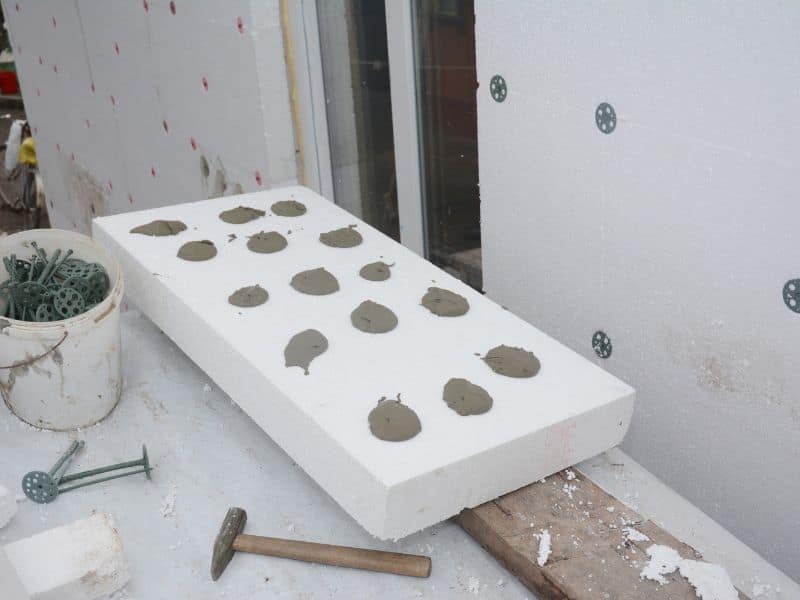 Rigid polystyrene insulation is attached to an adobe wall using blobs of adhesive and plastic cap nails.