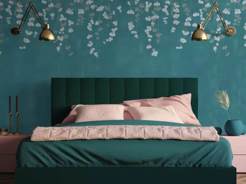 A bedroom in an adobe house with blue walls and green and pink furniture. Flowers are painted on the wall above the bed, and brass light fittings extend over the bed.