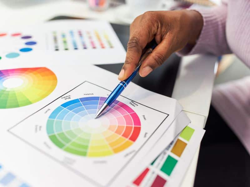 An interior designer points to a color wheel's center with a blue ballpoint pen. The wheel includes all the colors on the visible spectrum.