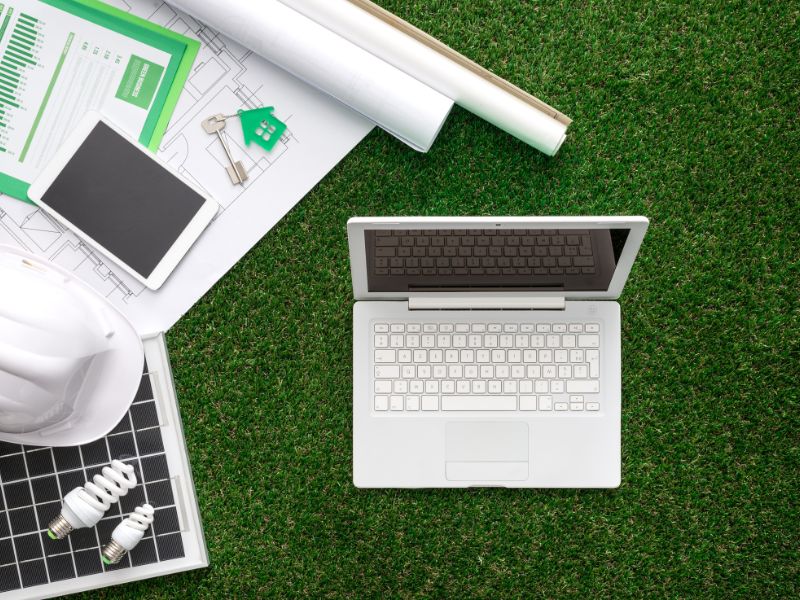 Sustainable building plans laid out on green grass with a key on a green house keyring, a small laptop, a tablet computer, energy-saving lightbulbs, and a white hard hat.