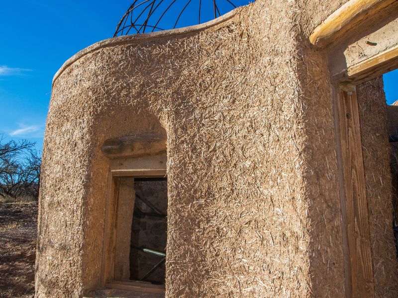 A cob house is under construction. The roof has not yet been built, but openings for the door and windows are in place with wooden lintels.