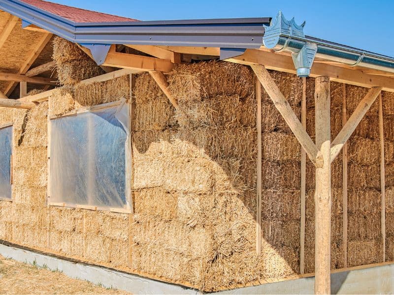 A straw bale house is under construction. The construction method is non-load bearing, with the weight of the roof being supported by wooden posts. Openings for windows are formed and covered with plastic sheeting as a temporary measure.