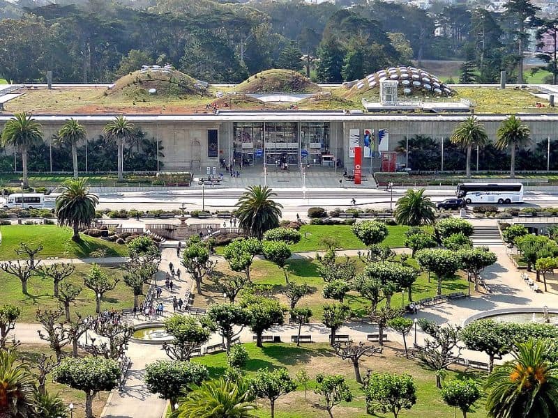 A photograph of the outside of the California Academy of Sciences and its grounds. It has a green roof and an ornamental park area with many trees and paths.