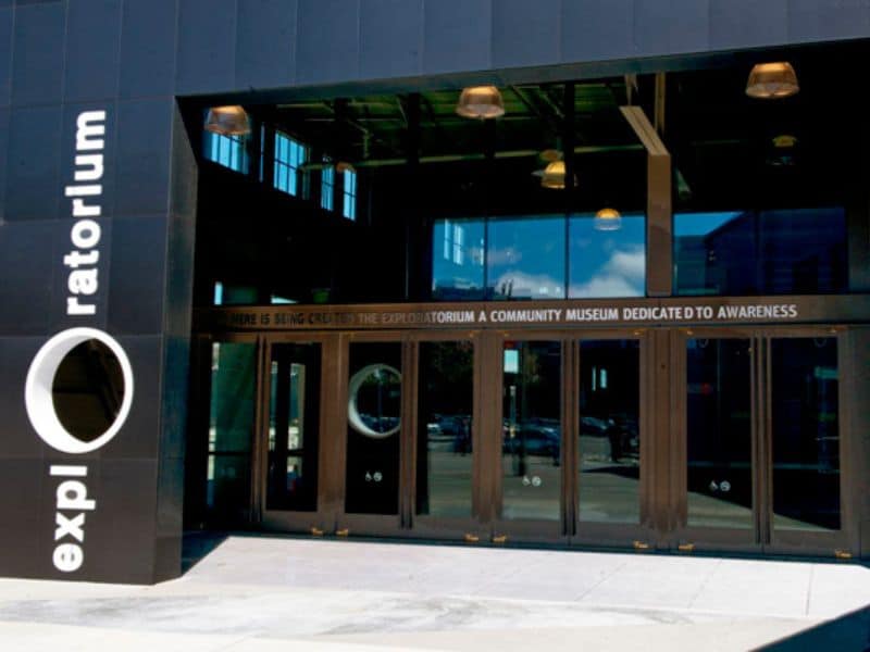 A photograph of the front entrance of the Exploratorium. The building is glass-fronted with stone paving outside.