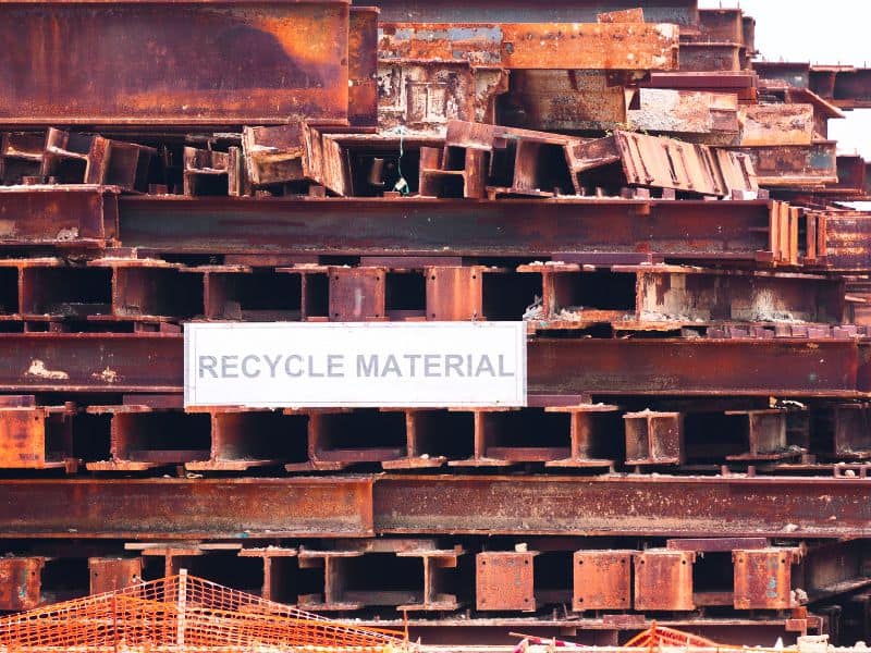 A stack of rusty orange steel members is ready for recycling. A sign states, "RECYCLE MATERIAL."