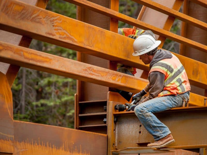 A steelworker connecting steel members on a bridge using bolts. The worker wears a hard hat and high visibility tabard.