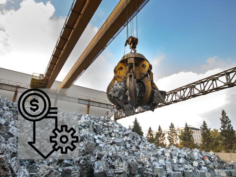 A large gantry-mounted mechanical grab lifts steel scrap for recycling. In the lower left of the image is a graphic showing a dollar sign with a downward pointing arrow.