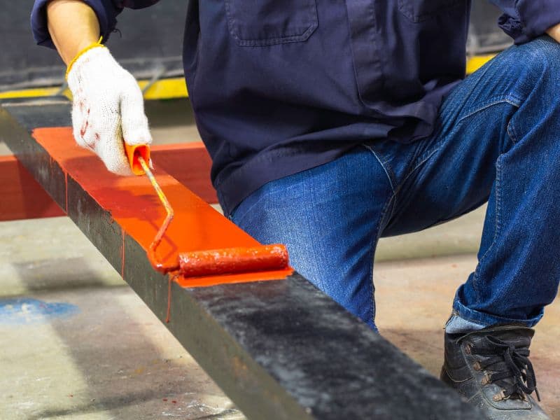 Using a roller, a worker kneels to paint a layer of red primer onto a steel beam. He wears white protective gloves, blue jeans, a shirt, and black work boots.