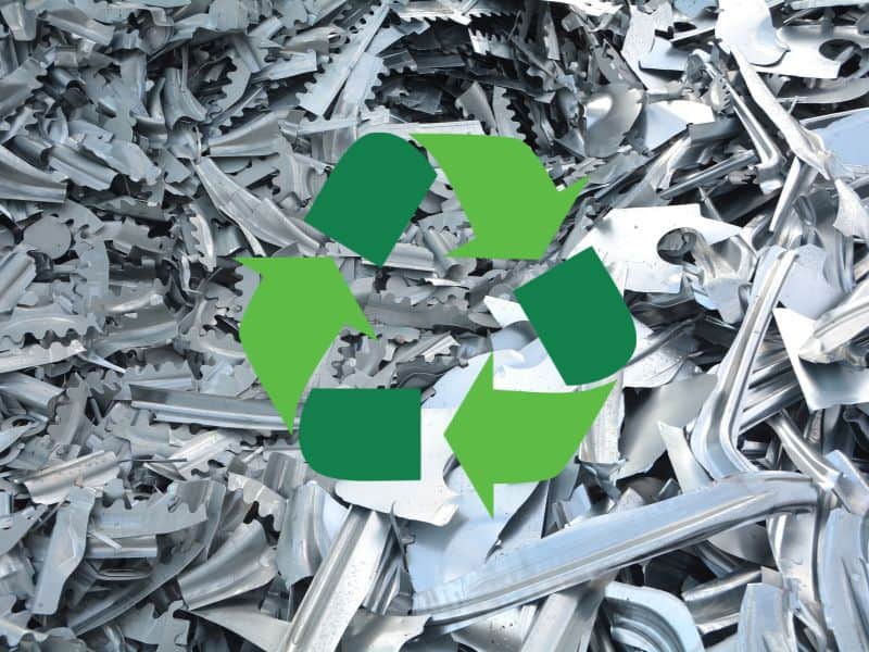 A photograph of mixed steel scrap, comprising off-cuts of various shapes and sizes. Overlain in the center of the image is a recycling symbol formed from three circular triangular arrows.