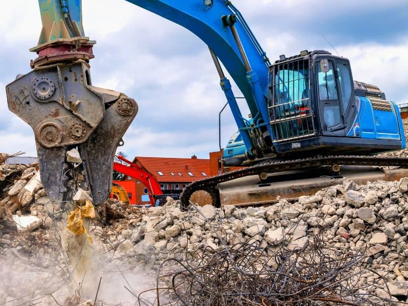 A large blue, tracked vehicle with a hydraulic grab separates and sorts through demolition waste on a demolition site to segregate steel rebar for recycling.