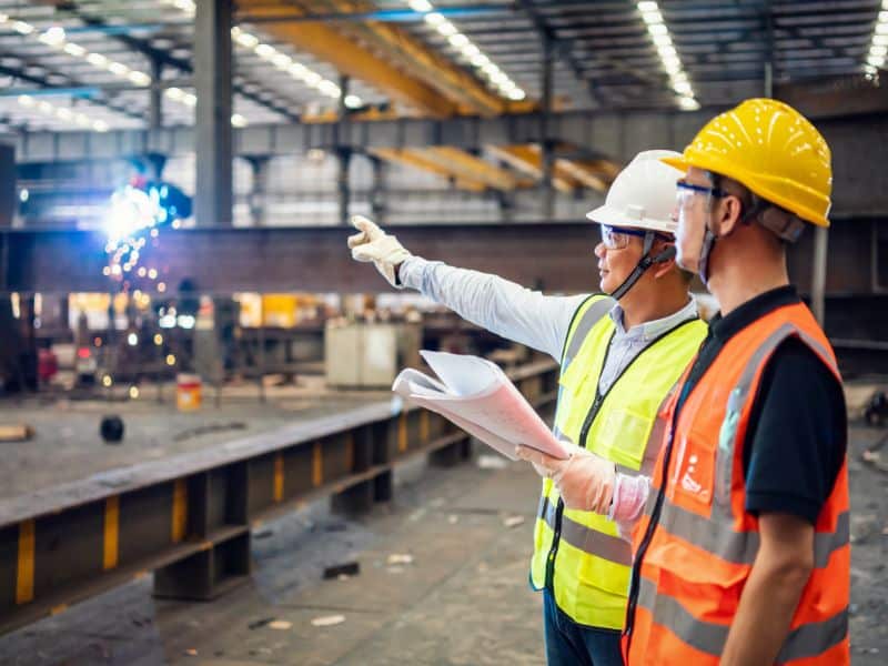 Two workers wearing hard hats, protective eye glasses, gloves and high visibility tabards discuss specifications of steelwork in a large warehouse.