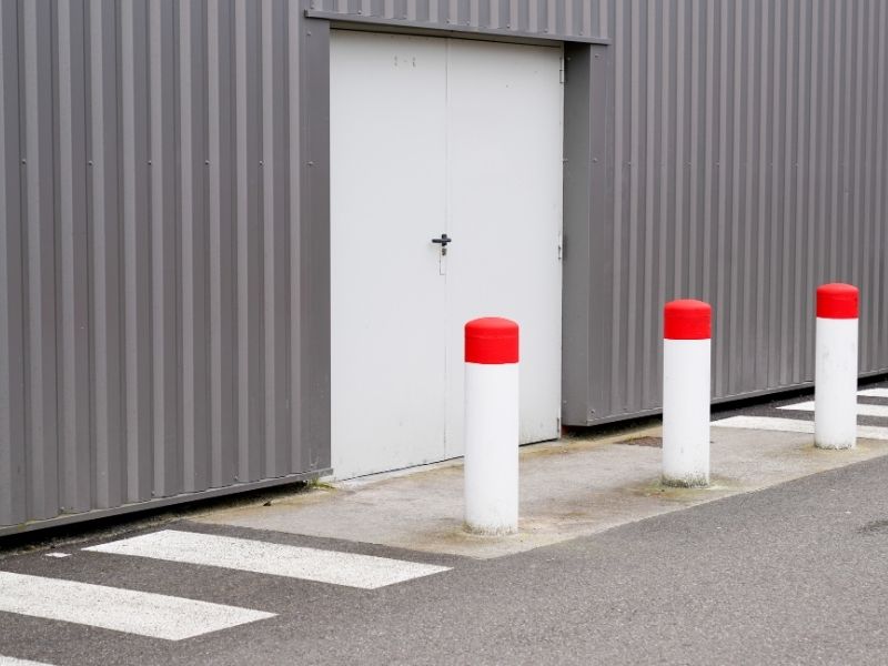 The outside of a warehouse with steel cladding on the walls. Large white double doors, protective bollards are outside the doors and a striped walkway is marked on the tarmac.