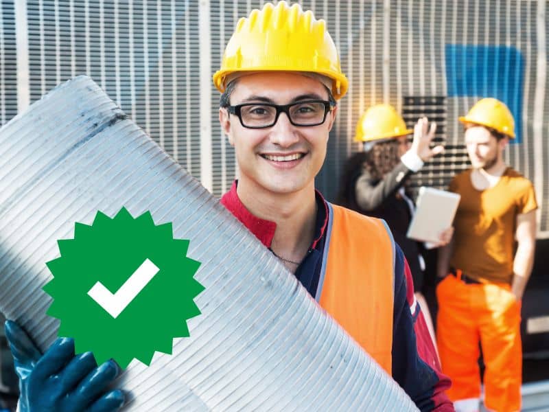 A worker wearing glasses, a yellow hard hat, an orange high-visibility tabard, and blue protective gloves carries a section of stainless steel flue. A green circle with a serrated edge and a white tick is in the bottom-left of the image. In the background on the right are two workers in a training session.