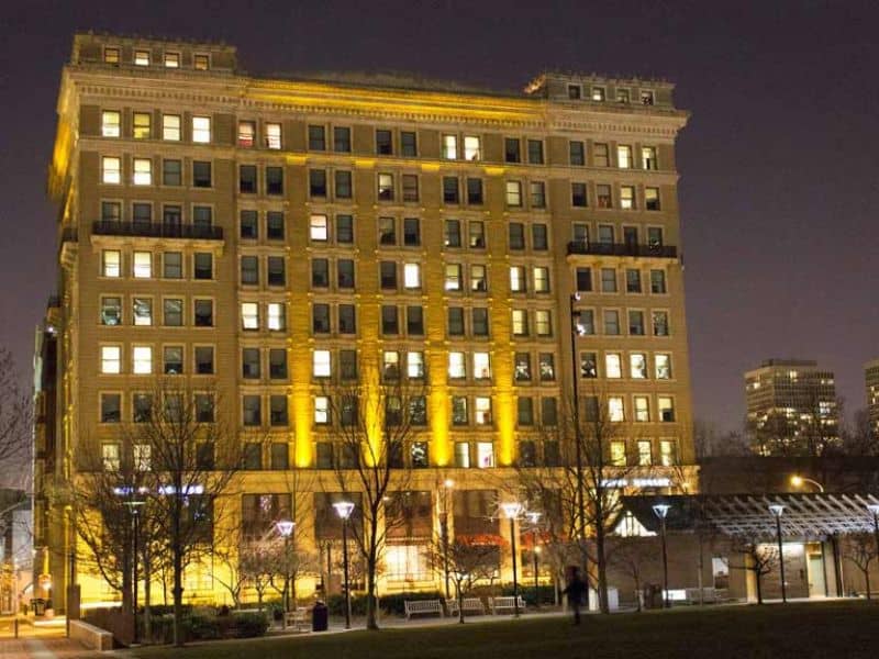 The Kimpton Hotel Monaco is seen at night. The rooms have lights on, and the lobby area is brightly lit. The stone façade has regularly spaced windows and balconies halfway up. Elaborate cornices overhang the top of the building.